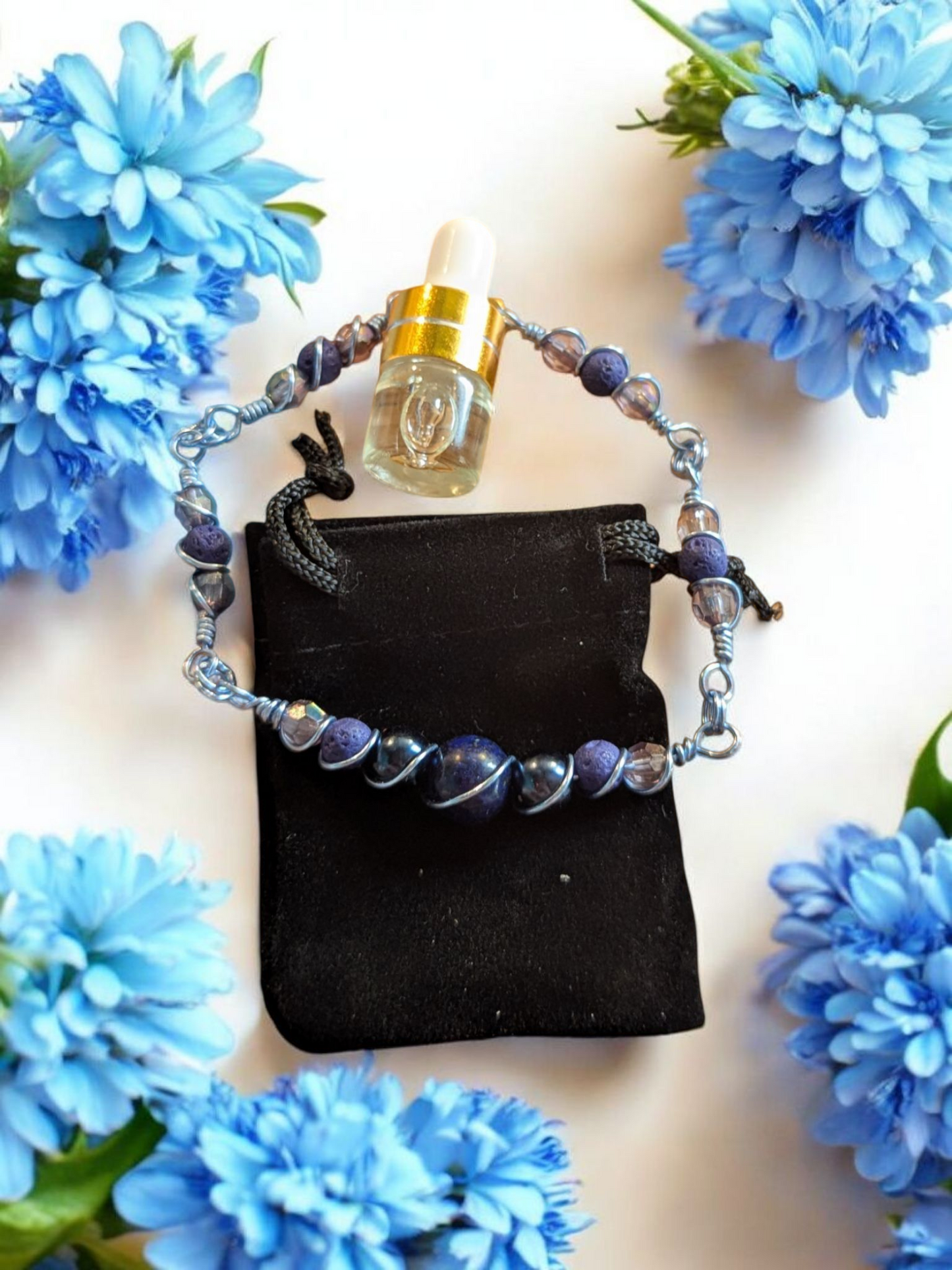 Handcrafted lapis lazuli diffuser bracelet with dark blue lava beads, resin pearls, and iridescent gray accents wrapped in light blue hypoallergenic wire, displayed on a black velvet pouch surrounded by blue flowers.