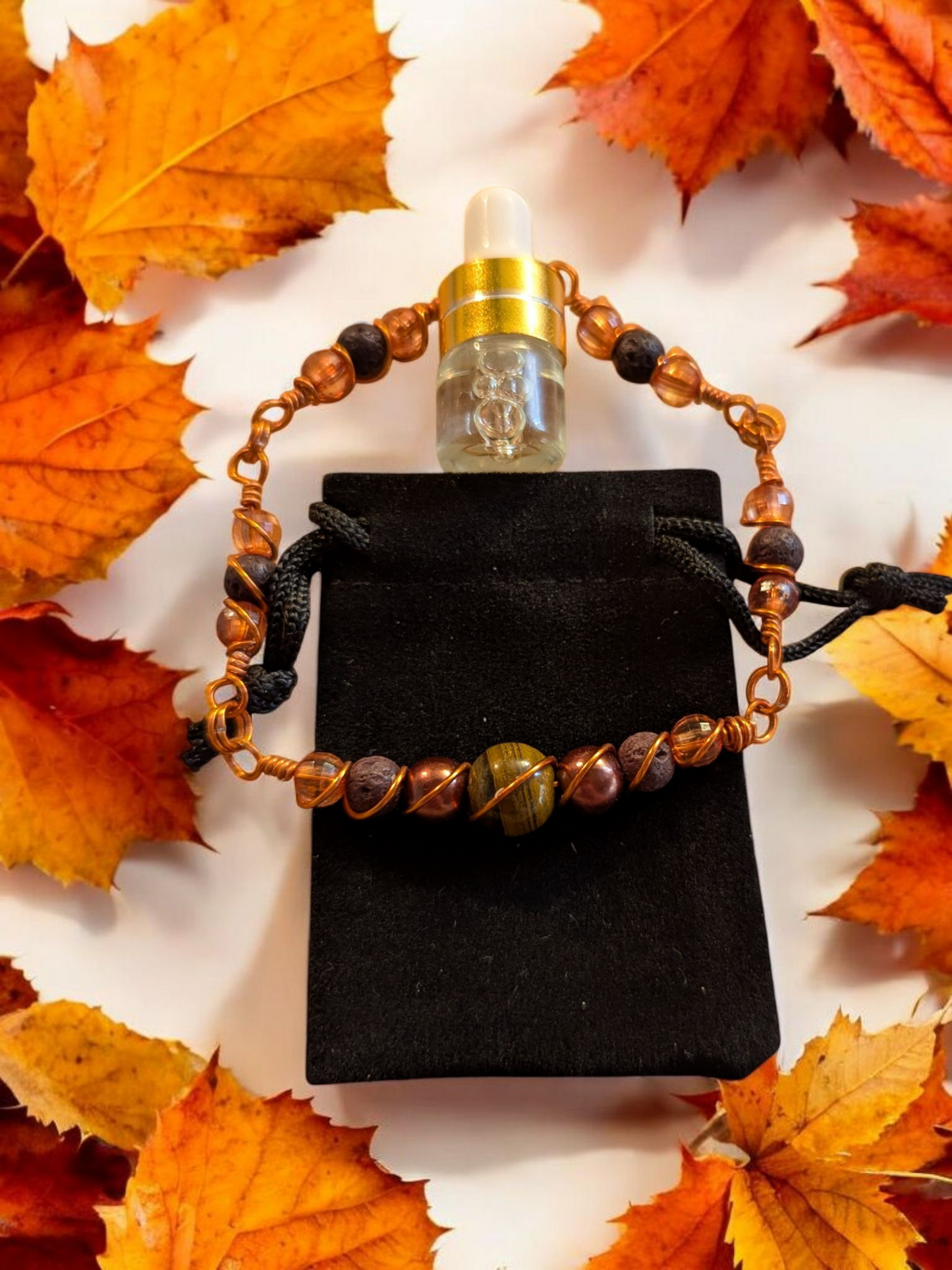 Handcrafted Tiger’s Eye diffuser bracelet with black lava stones, amber resin beads, and copper hypoallergenic wire, displayed on a black velvet pouch surrounded by autumn leaves.