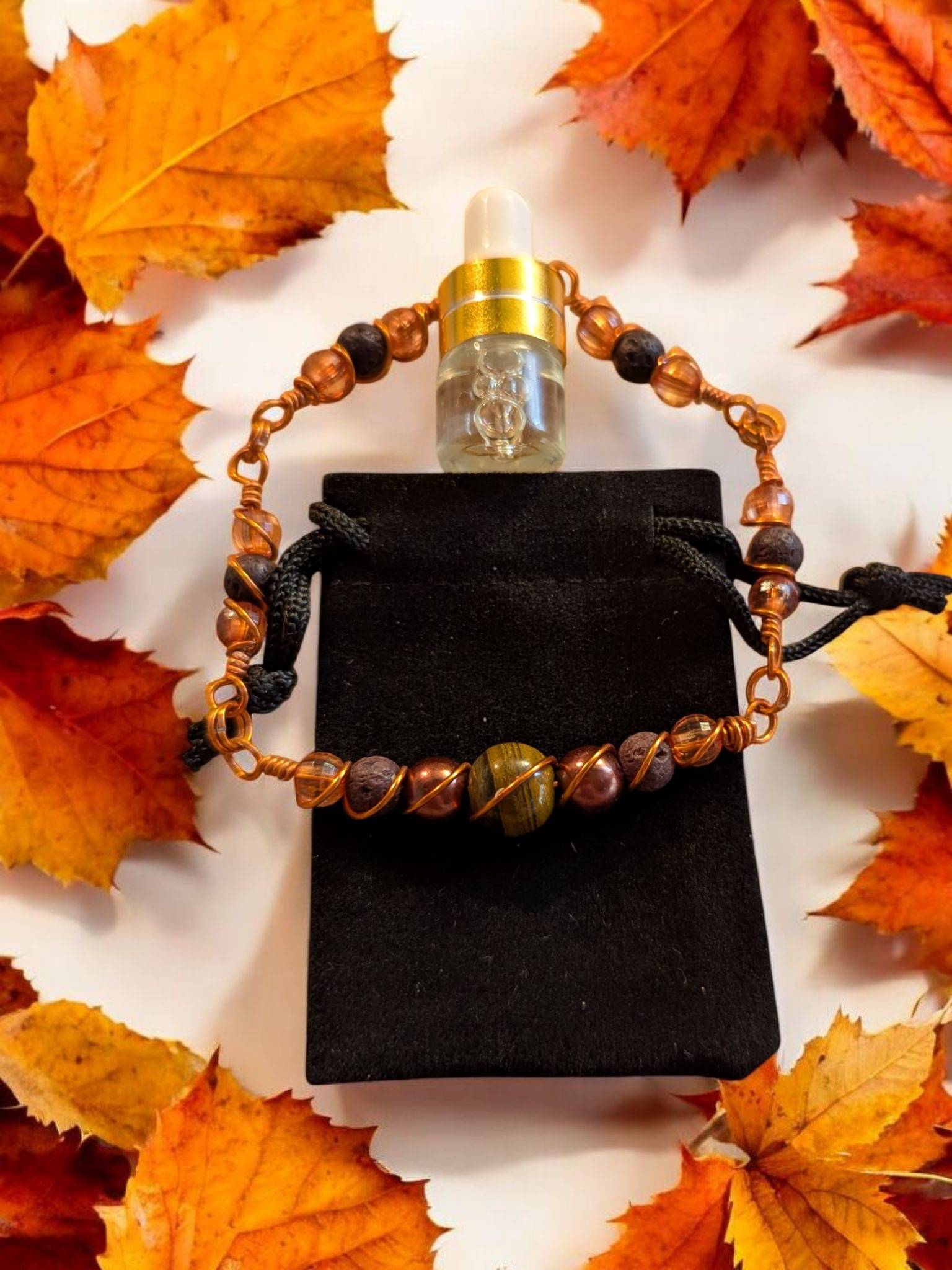 Handcrafted Tiger’s Eye diffuser bracelet with black lava stones, amber resin beads, and copper hypoallergenic wire, displayed on a black velvet pouch surrounded by autumn leaves.