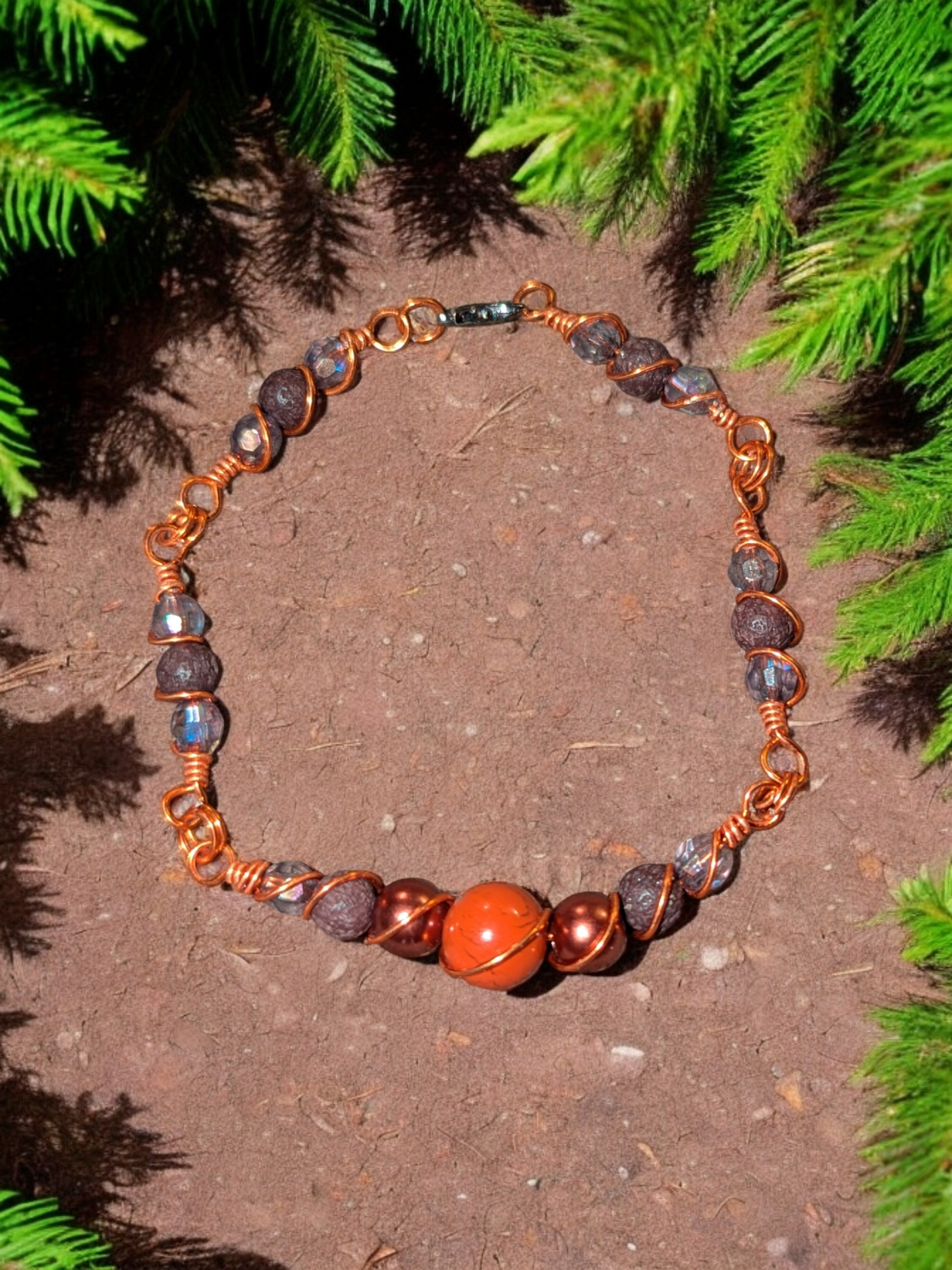Handcrafted red agate diffuser bracelet with bronze resin pearls, dark brown lava beads, iridescent rondel accents, and bright copper hypoallergenic wire, displayed on earthy ground surrounded by evergreen branches.