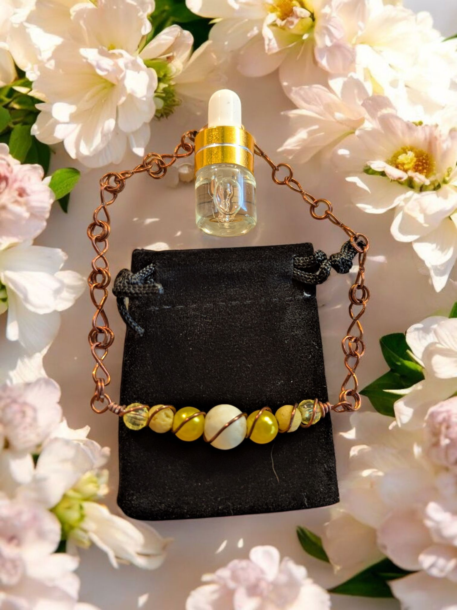 Handcrafted yellow jade diffuser bracelet with yellow lava beads, resin pearls, and copper hypoallergenic wire with infinity links, displayed on a black velvet pouch surrounded by white flowers.