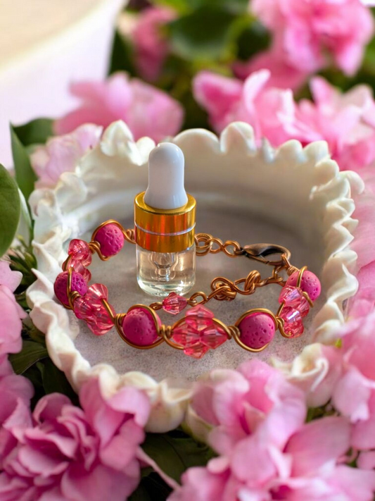 Handcrafted adjustable cuff diffuser bracelet with pink lava stones and pink resin bicone beads wrapped in copper-colored hypoallergenic wire, displayed in a white ceramic dish surrounded by pink flowers.