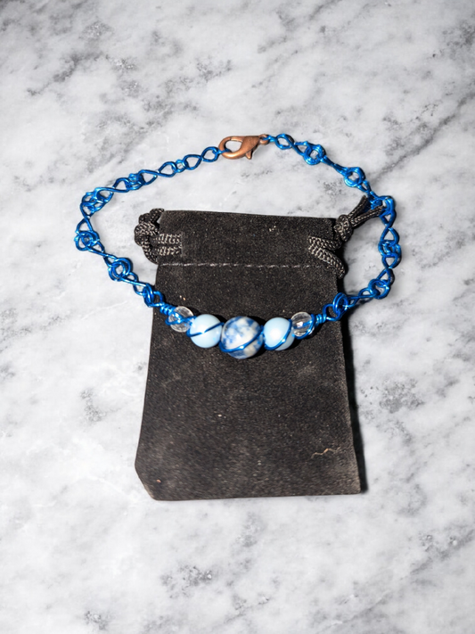 Frostbite Halo Eternity Bracelet with blue-colored wire-wrapped marbled blue and white sodalite stone beads, resin pearls and clear resin beads displayed on a black pouch on a marble background.