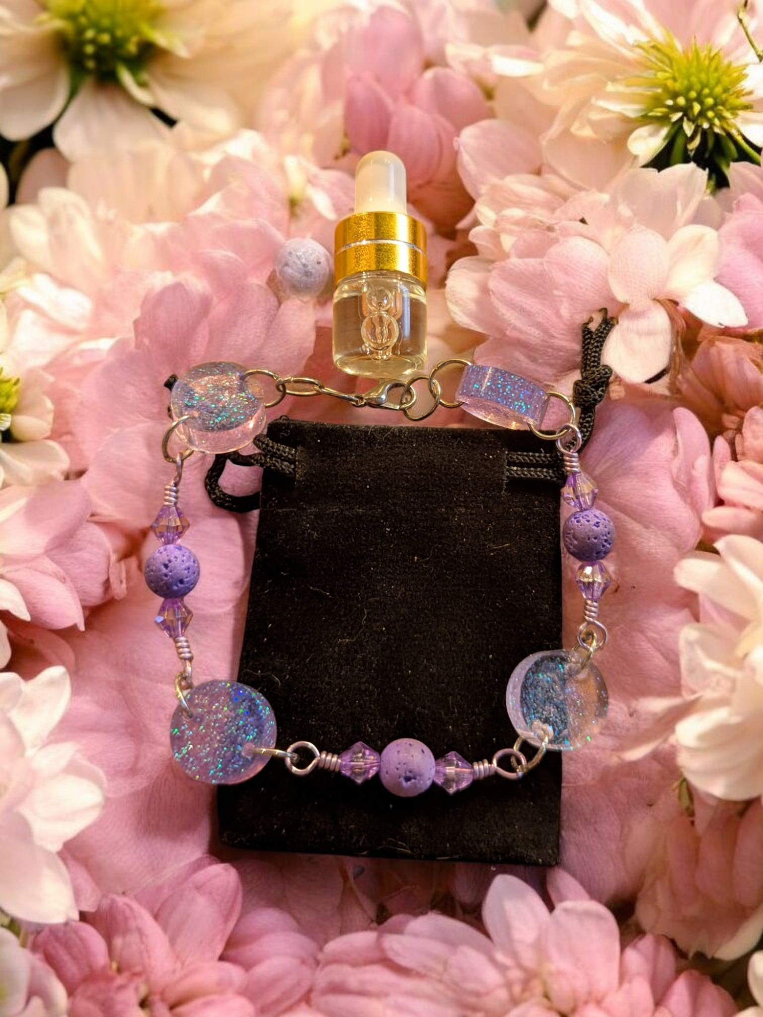 Handcrafted lavender diffuser bracelet with clear resin glitter discs, lavender lava beads, and lavender hypoallergenic wire displayed on a black velvet pouch surrounded by pink and white flowers.