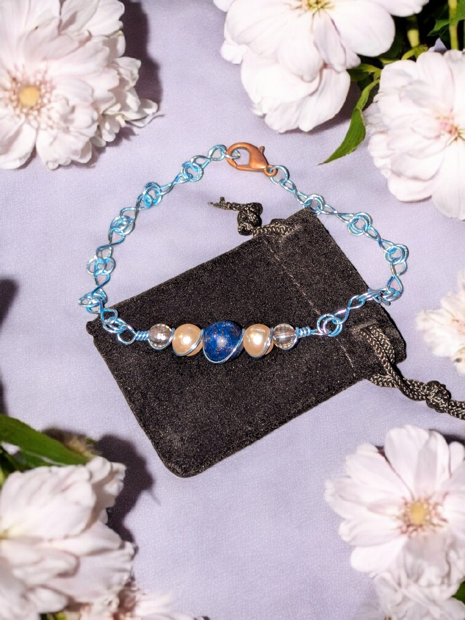 Blue wire love-knot bracelet with pearls and a lapis lazuli centerpiece displayed on a black pouch surrounded by white flowers.