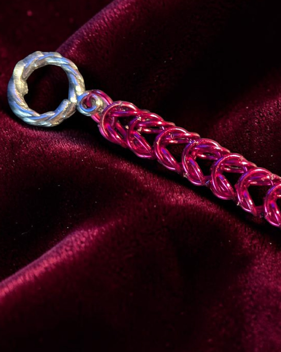A close-up macro of the Brikk box weave chainmail bracelet in Magenta, its bright pink-magenta aluminum rings densely interlocked in the structural box weave pattern across deep burgundy velvet, with the silver toggle ring clasp visible at the left.