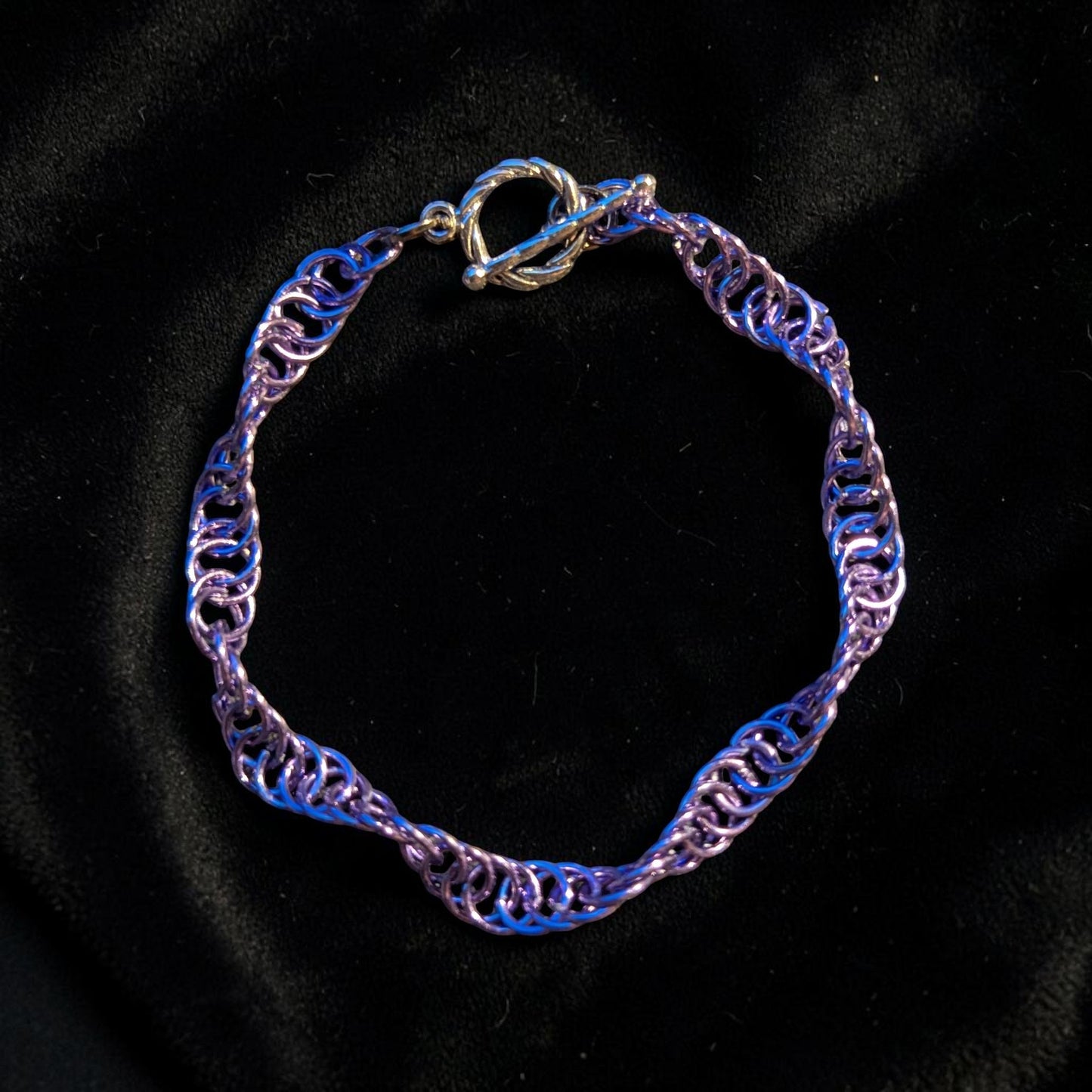 The Snerill spiral 4-in-1 chainmail bracelet in Lavender laid in a circle on a dark gray surface with a purple light cast, its soft pinkish-lavender aluminum rings twisting in a continuous helix and finished with a silver toggle clasp at the top.