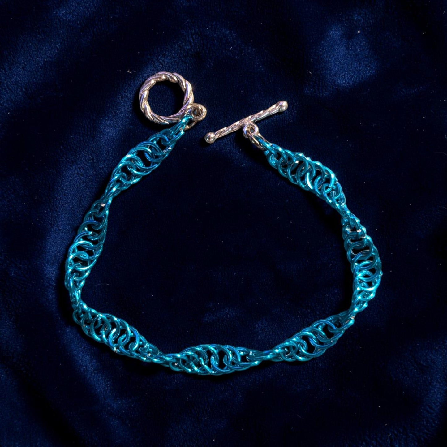 The Snerill spiral 4-in-1 chainmail bracelet in Aqua laid open on deep blue velvet, its bright turquoise aluminum rings twisting in a continuous helix with the silver toggle clasp unfastened at the top.