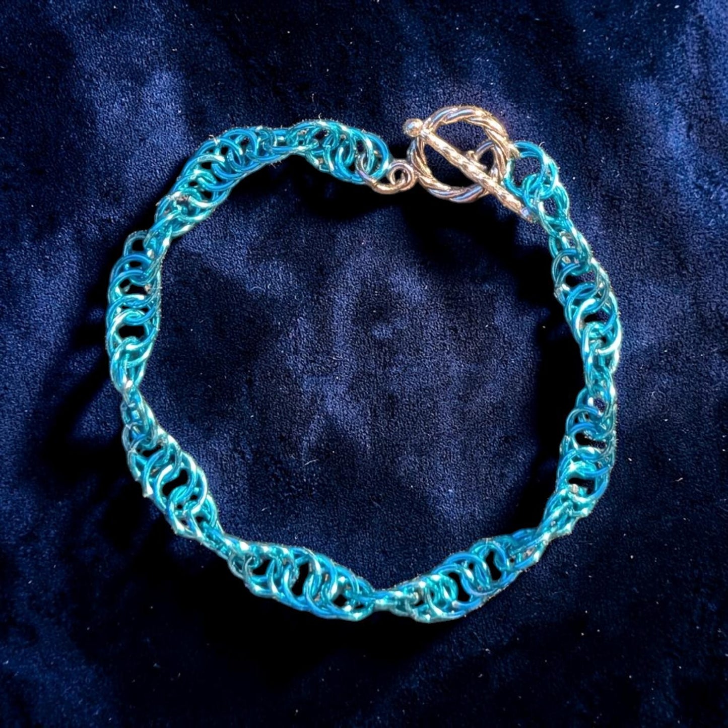 The Snerill spiral 4-in-1 chainmail bracelet in Pale-Aqua laid in a circle on dark blue velvet, its soft turquoise aluminum rings twisting in a continuous helix and finished with a silver toggle clasp at the top.