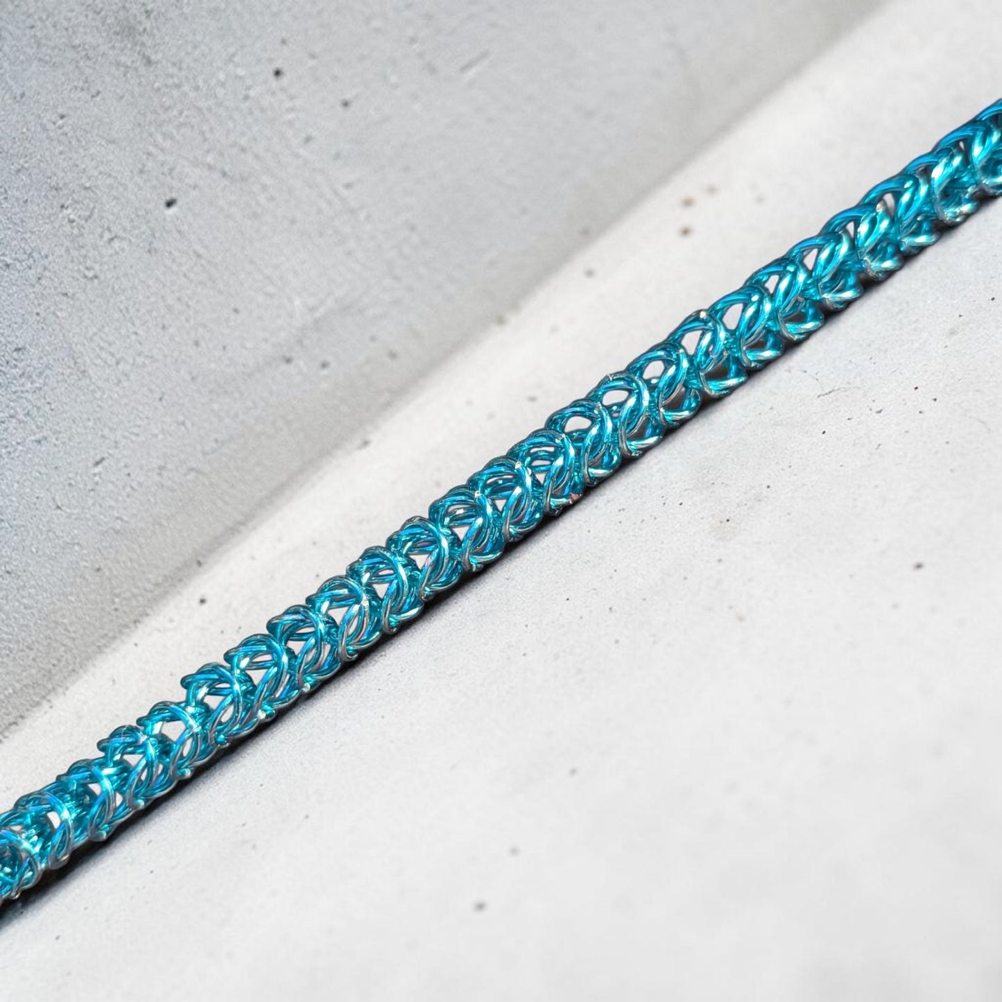 A close-up macro of the Brikk box weave chainmail bracelet in Pale-Aqua, its bright sky-blue aluminum rings densely interlocked in the structural box weave pattern laid diagonally across a pale concrete surface.