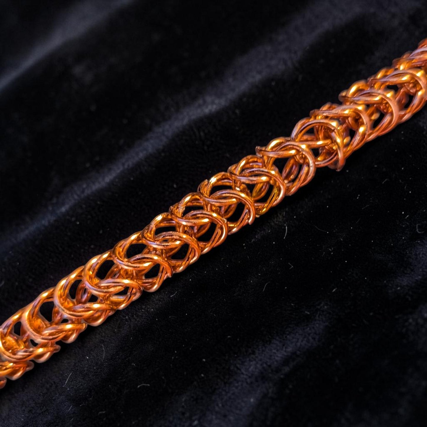  close-up macro of the Brikk box weave chainmail bracelet in Rose-Gold, its aluminum rings densely interlocked in the structural box weave pattern across black velvet, lit to give the rose-gold finish a warm orange-copper tone.