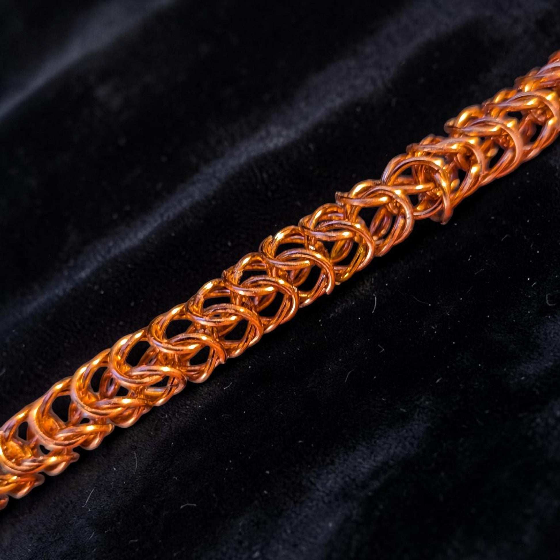  close-up macro of the Brikk box weave chainmail bracelet in Rose-Gold, its aluminum rings densely interlocked in the structural box weave pattern across black velvet, lit to give the rose-gold finish a warm orange-copper tone.