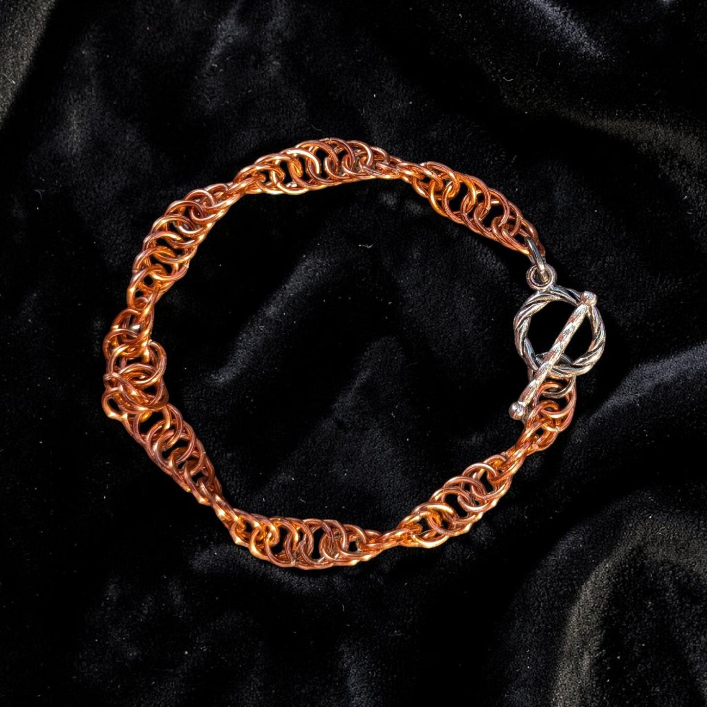 The Snerill spiral 4-in-1 chainmail bracelet in Rose-Gold laid in a circle on black velvet, its warm peachy-copper aluminum rings twisting in a continuous helix and finished with a silver toggle clasp at the right.