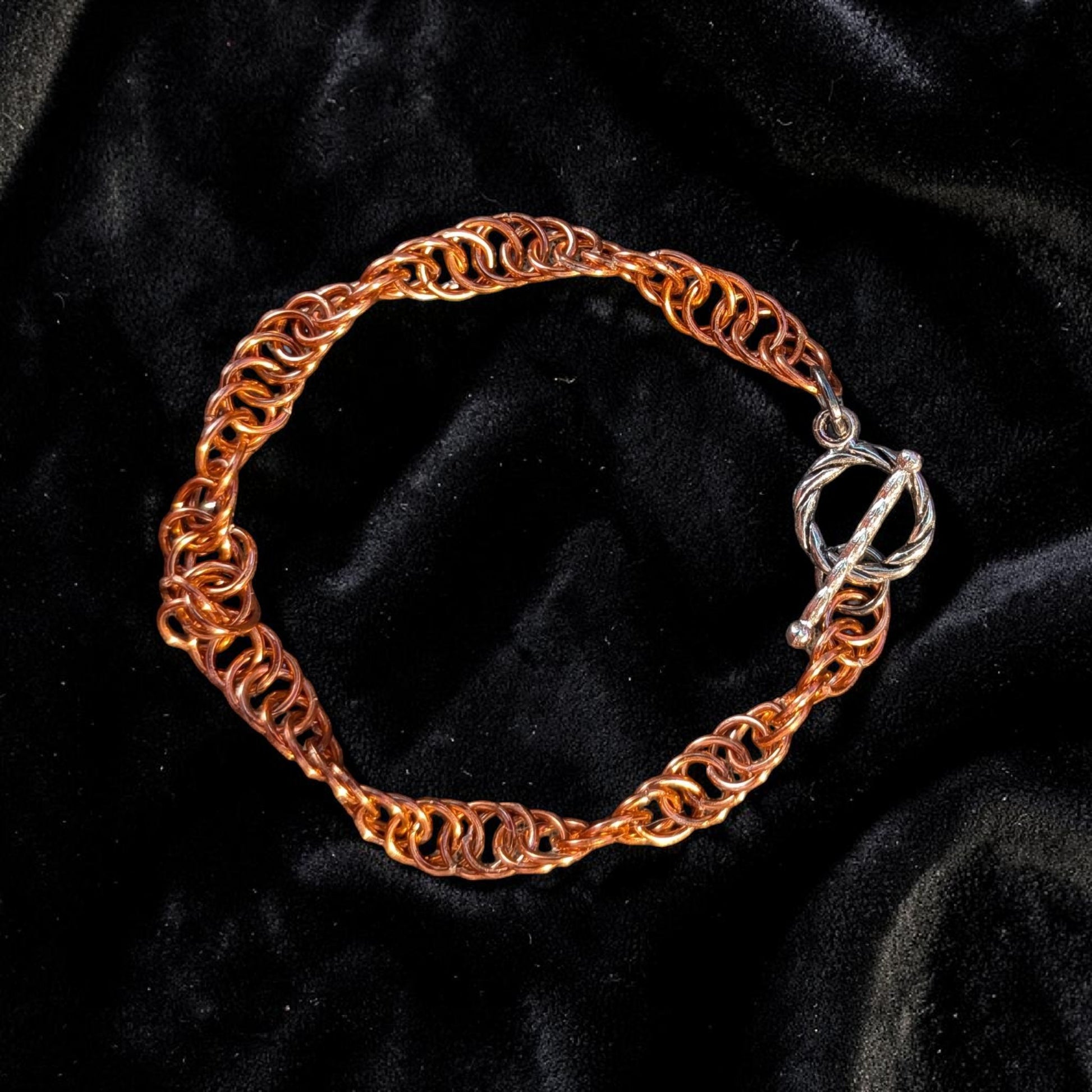 The Snerill spiral 4-in-1 chainmail bracelet in Rose-Gold laid in a circle on black velvet, its warm peachy-copper aluminum rings twisting in a continuous helix and finished with a silver toggle clasp at the right.
