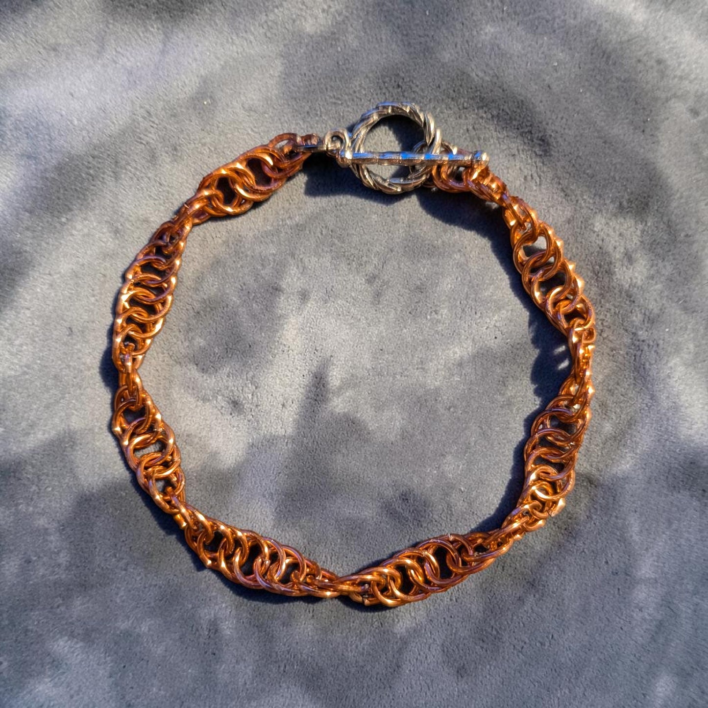 The Snerill spiral 4-in-1 chainmail bracelet in Copper laid in a circle on textured gray concrete, its bright orange-copper aluminum rings twisting in a continuous helix and finished with a silver toggle clasp at the top.