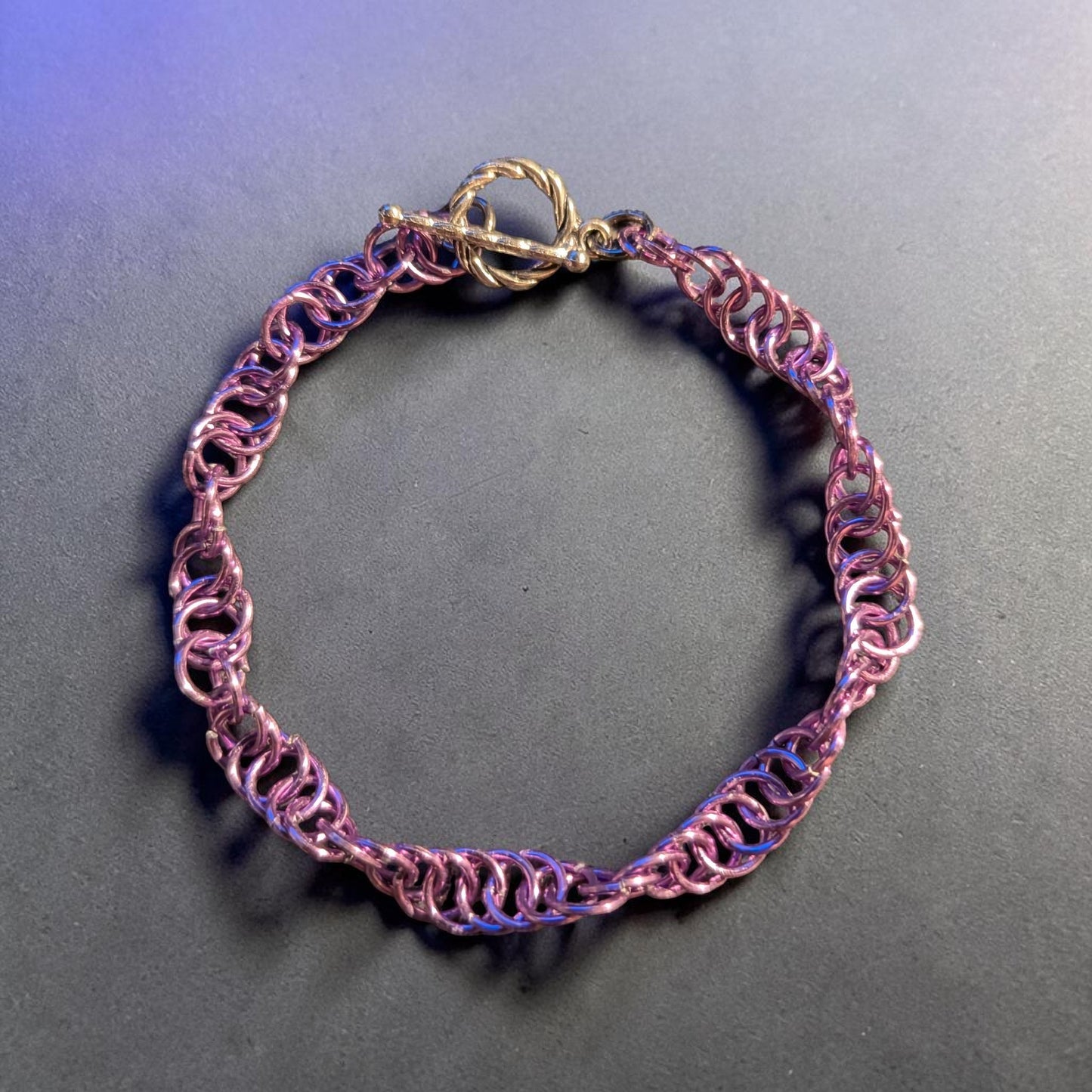 The Snerill spiral 4-in-1 chainmail bracelet in Lavender laid in a circle on a dark gray surface with a purple light cast, its soft pinkish-lavender aluminum rings twisting in a continuous helix and finished with a silver toggle clasp at the top.