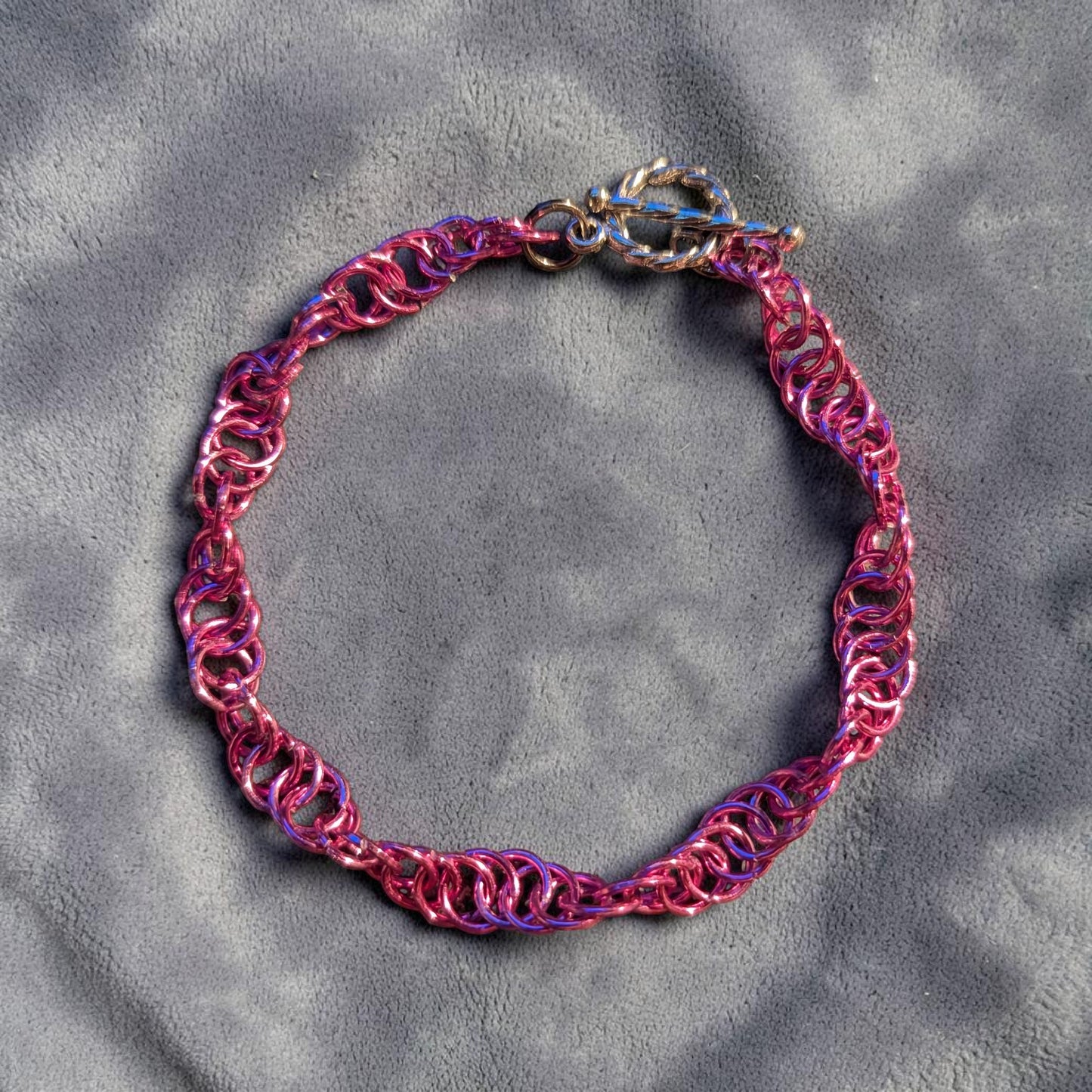 The Snerill spiral 4-in-1 chainmail bracelet in Rose-Pink laid in a circle on soft gray velvet, its bright pink aluminum rings twisting in a continuous helix and finished with a silver toggle clasp at the top.