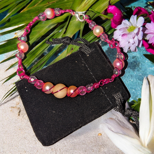 orbital wire-wrapped bracelet with warm jade and rose-toned beads and hot pink colored wire displayed on a black pouch near tropical flowers.