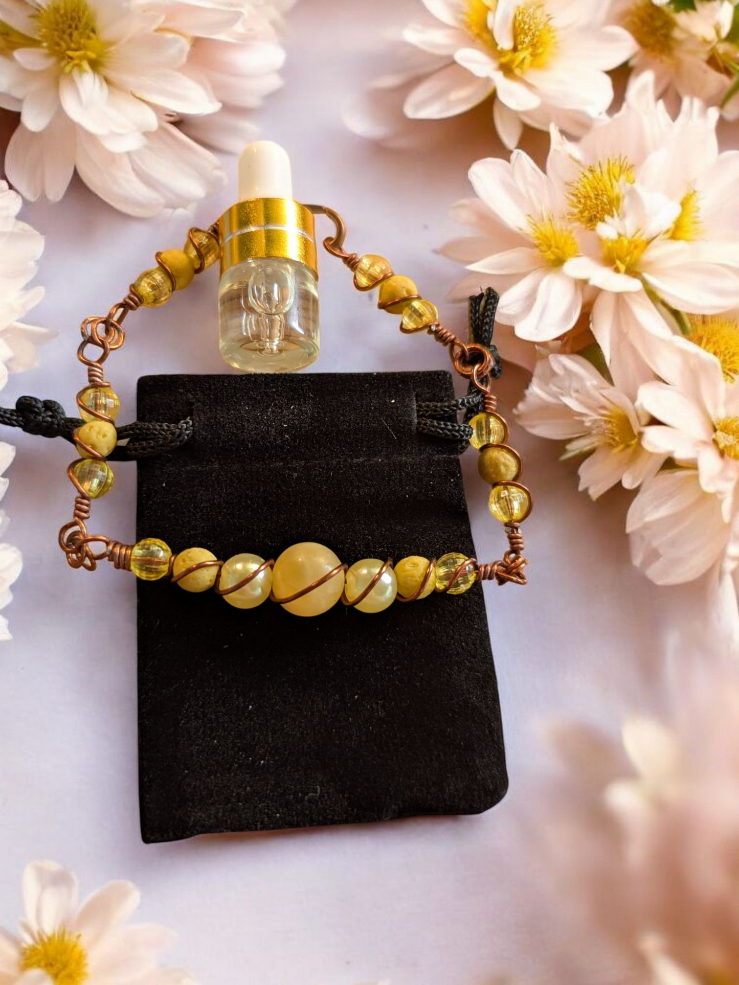 Handcrafted yellow jade diffuser bracelet with yellow lava beads, resin pearls, and rondel accents wrapped in dark copper hypoallergenic wire, displayed on a black velvet pouch surrounded by pale flowers.