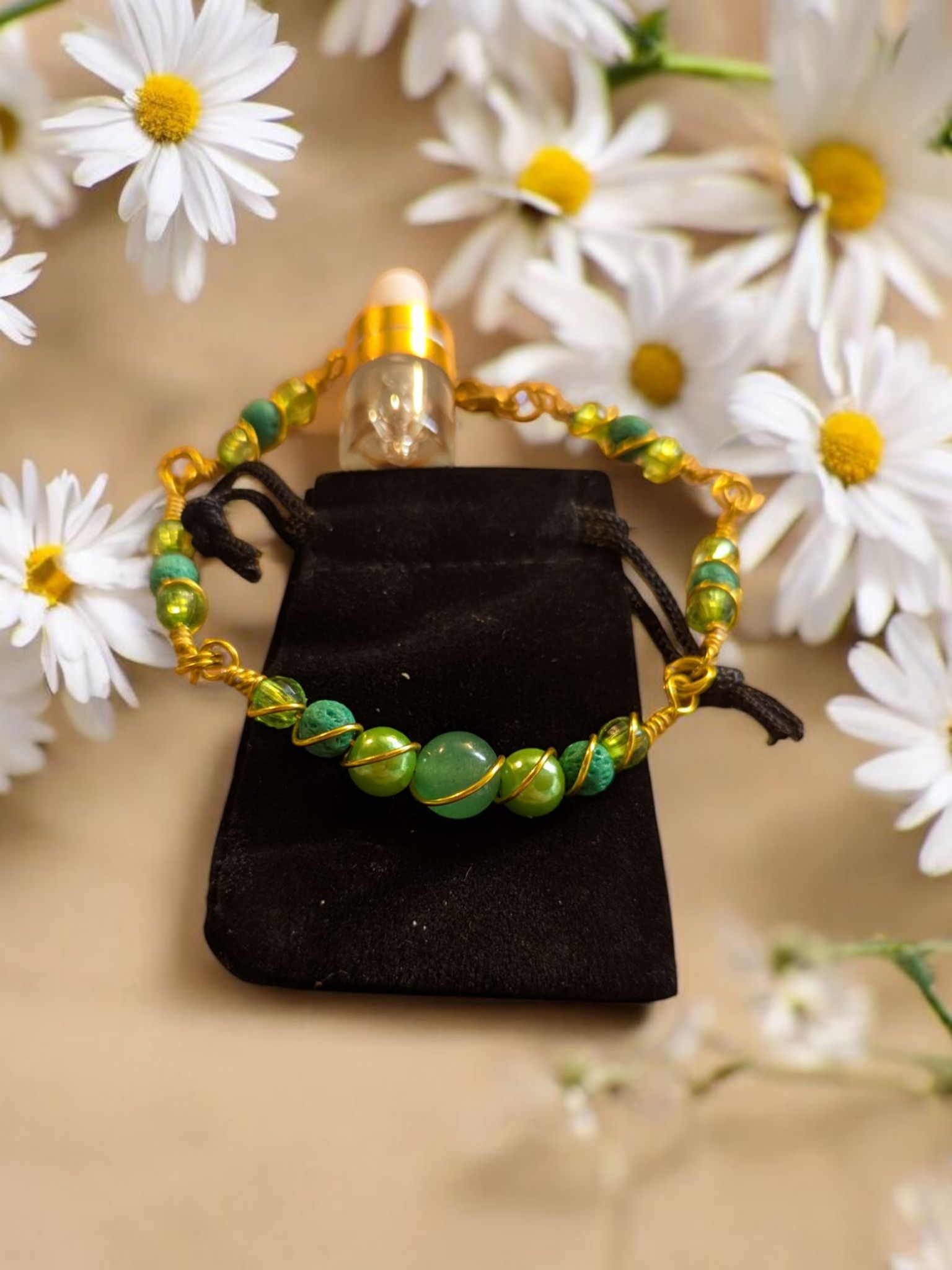 Handcrafted green aventurine diffuser bracelet with green lava stones, light green-gold resin pearls, and gold-colored hypoallergenic wire, displayed on a black velvet pouch surrounded by white daisies.