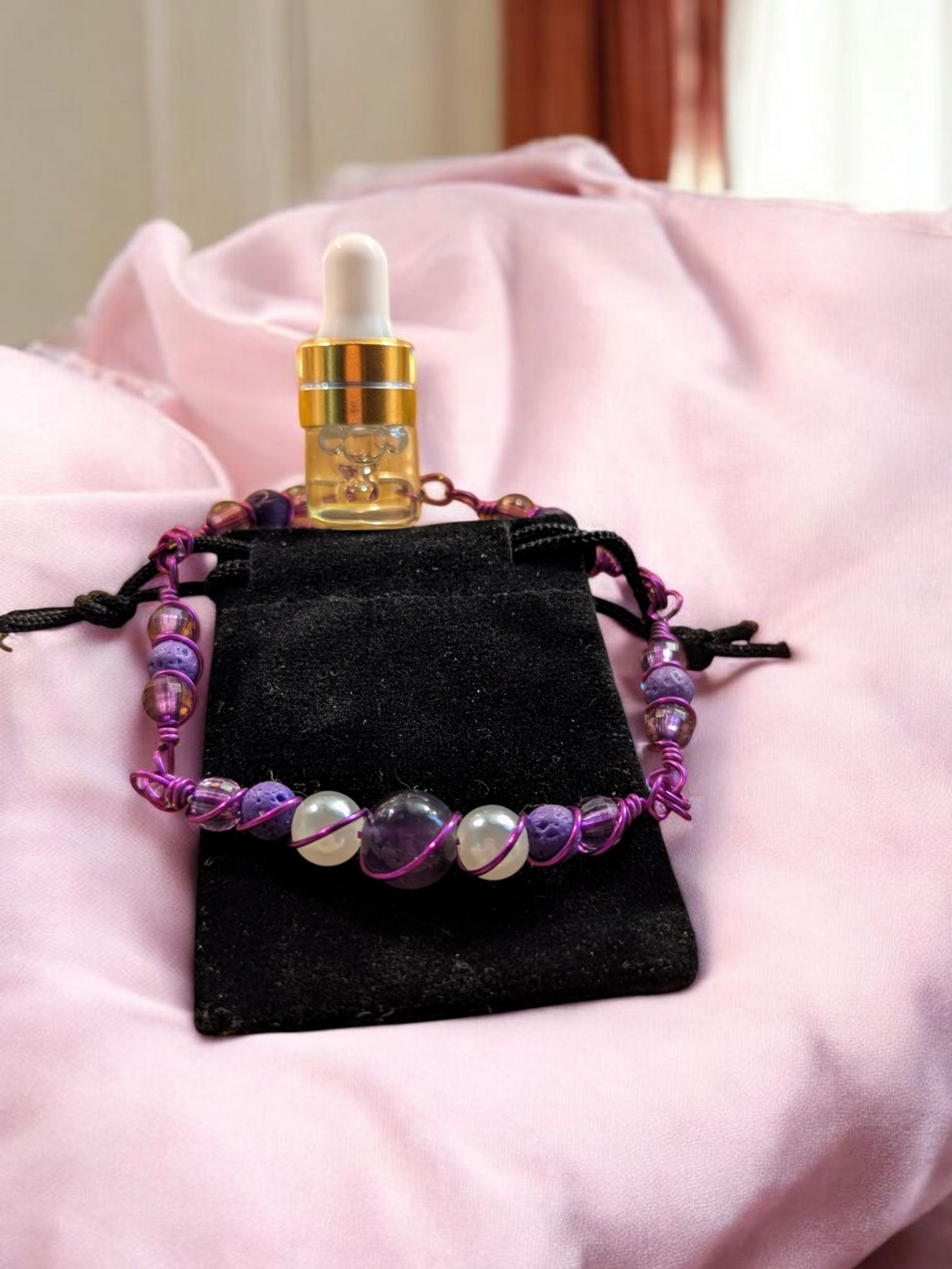 Product photo of the Violet Veil amethyst and lava diffuser link bracelet displayed on a black velvet pouch, with a small gold-capped glass dropper vial behind it, all resting on soft pink fabric in natural light.