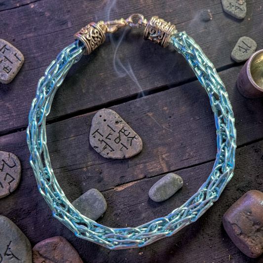 Unisex Viking braid bracelet made of shiny pale blue-silver woven wire arranged in a circle on dark wooden planks. The bracelet has ornate silver-toned end caps and a hook-and-ring clasp at the top. Smooth river stones surround it, including one flat stone with black rune-like markings. A faint wisp of smoke curls upward inside the bracelet circle.