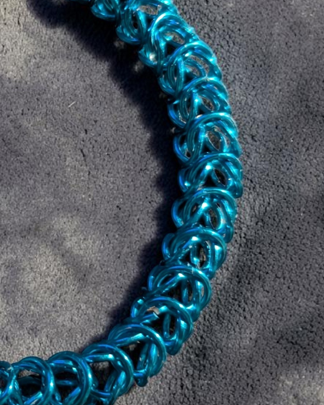 A close-up macro of the Brikk box weave chainmail bracelet in Aqua, its bright turquoise aluminum rings densely interlocked in the structural box weave pattern on a gray velvet surface in direct sunlight.