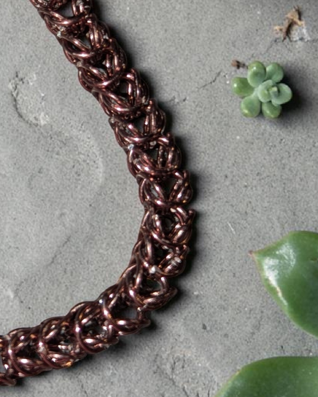 A close-up macro of the Brikk box weave chainmail bracelet in Antique-Bronze, its deep bronze-toned aluminum rings densely interlocked in the structural box weave pattern on a gray concrete surface with a small succulent and green leaves nearby.