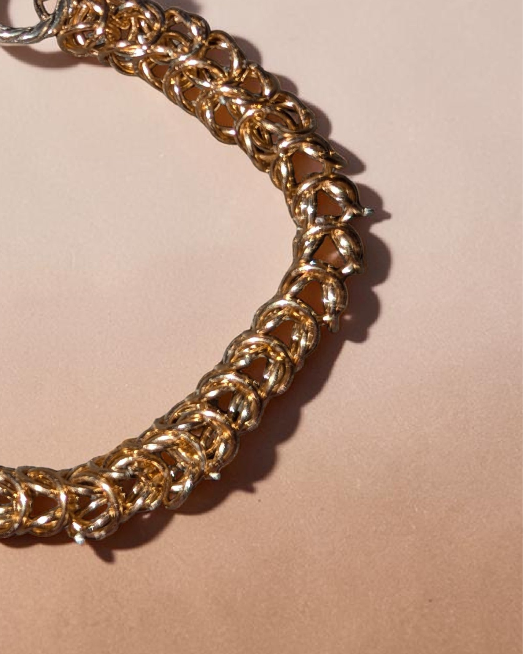 The Brikk box weave chainmail bracelet in Champagne laid in a circle on a soft peach-beige background, its warm pale-gold aluminum rings densely interlocked in the structural box weave pattern and finished with a silver toggle clasp at the top.