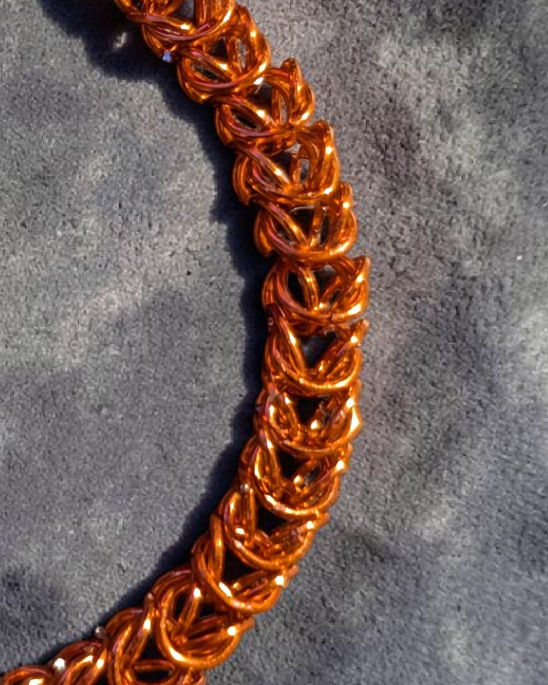 A close-up macro of the Brikk box weave chainmail bracelet in Copper, its bright orange-copper aluminum rings densely interlocked in the structural box weave pattern on a gray velvet surface in direct sunlight.