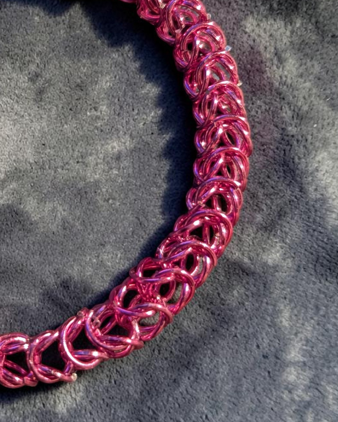 A close-up macro of the Brikk box weave chainmail bracelet in Rose-Pink, its bright pink aluminum rings densely interlocked in the structural box weave pattern across gray velvet in sunlight.
