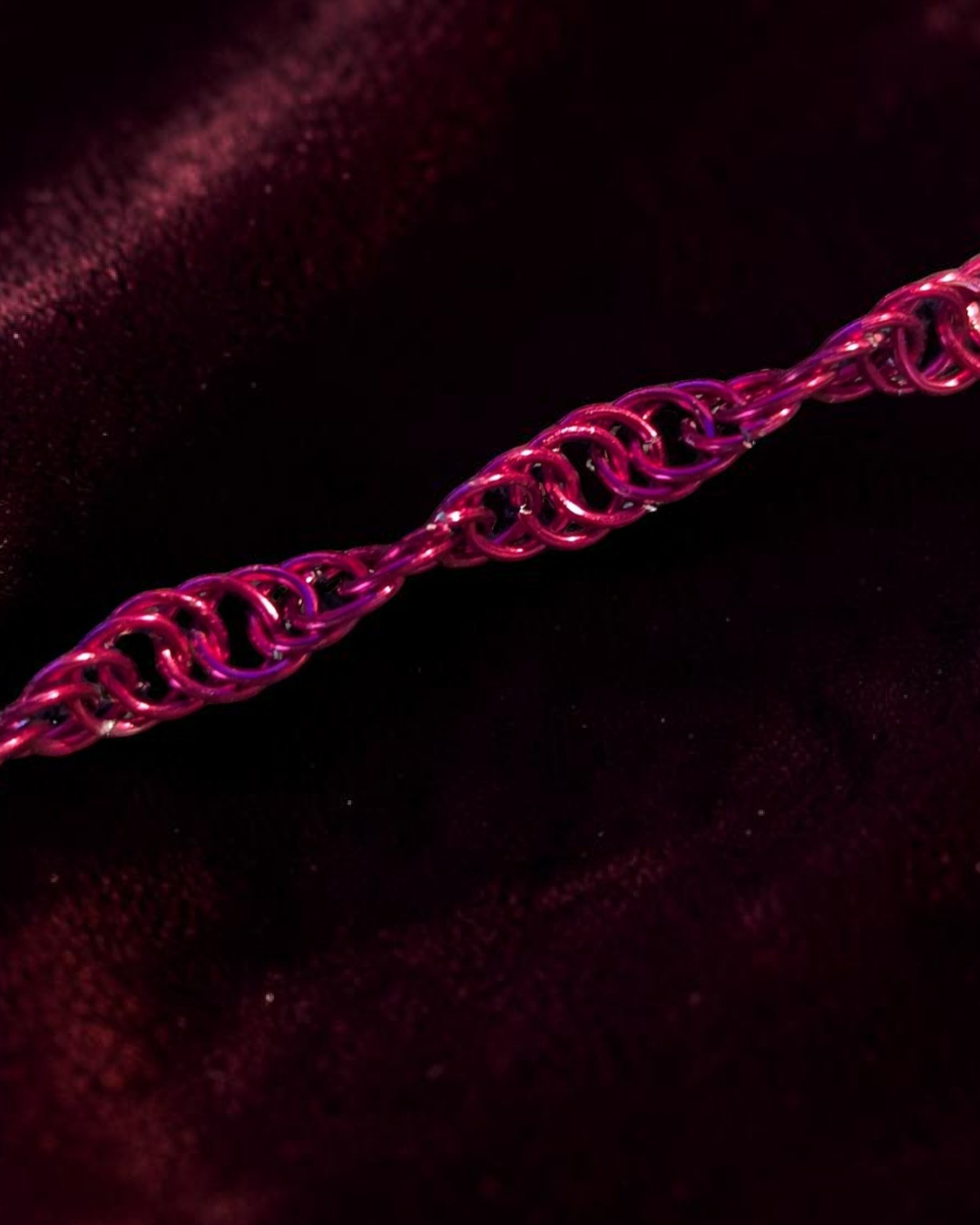 A close-up macro of the Snerill spiral 4-in-1 chainmail bracelet in Magenta, its bright pink-magenta aluminum rings twisting in a continuous helix across deep burgundy velvet catching a single highlight of light.