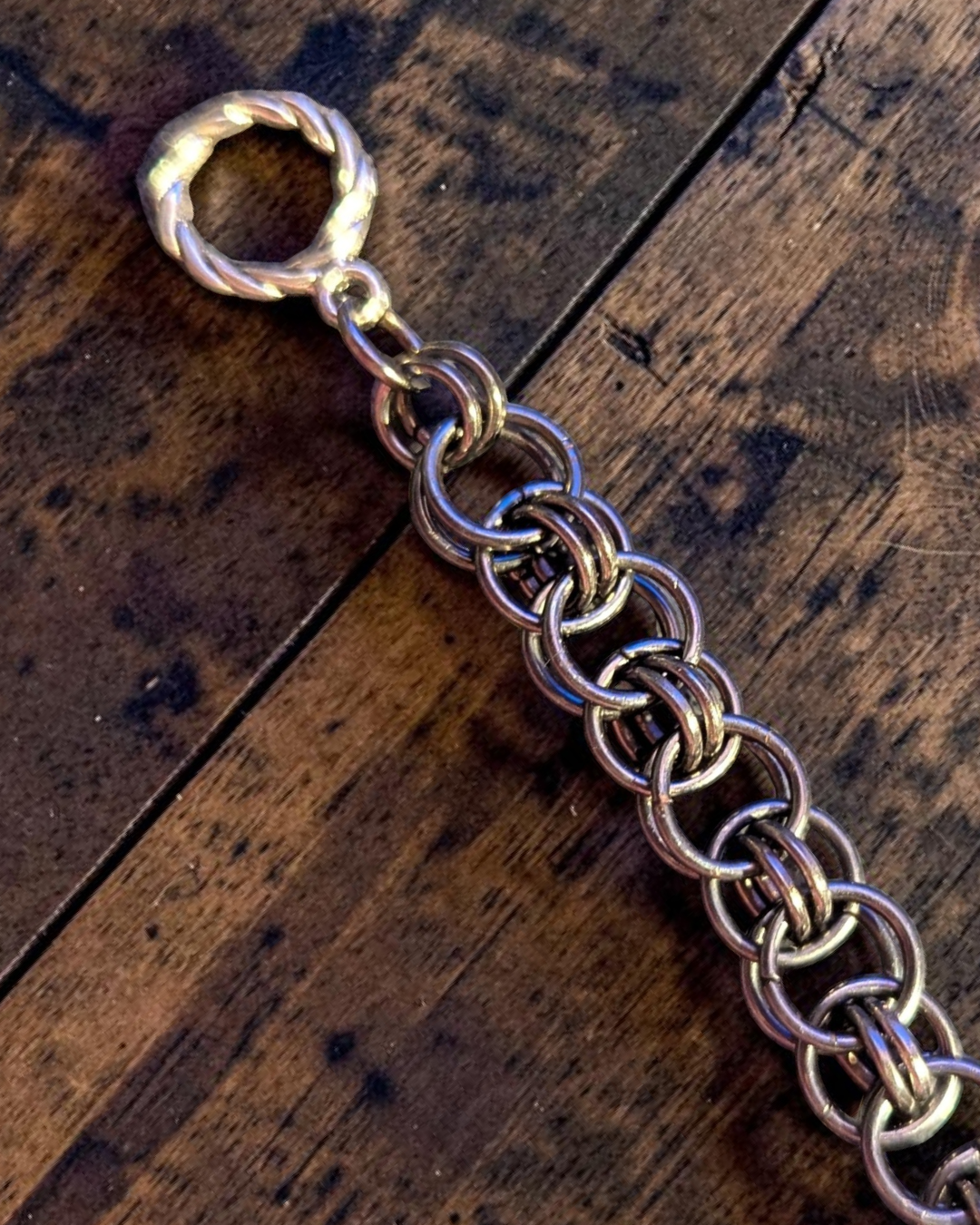 Close-up of Yalm, a Helm Chain bracelet laid diagonally on dark stained wood. The bracelet has interlocked gun-metal black rings with a hematite-like finish and a repeating chainmail pattern made from larger outer rings and smaller connecting rings. A twisted round clasp and connector rings are visible at one end, with the rest of the bracelet extending out of frame.